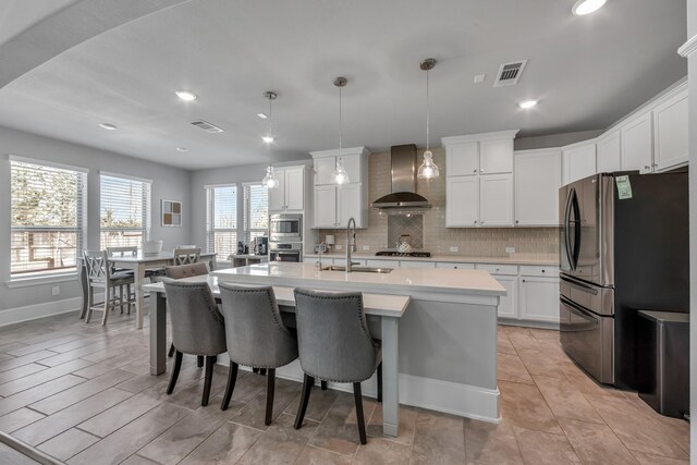 kitchen with sink, hanging light fixtures, stainless steel appliances, a center island with sink, and wall chimney exhaust hood