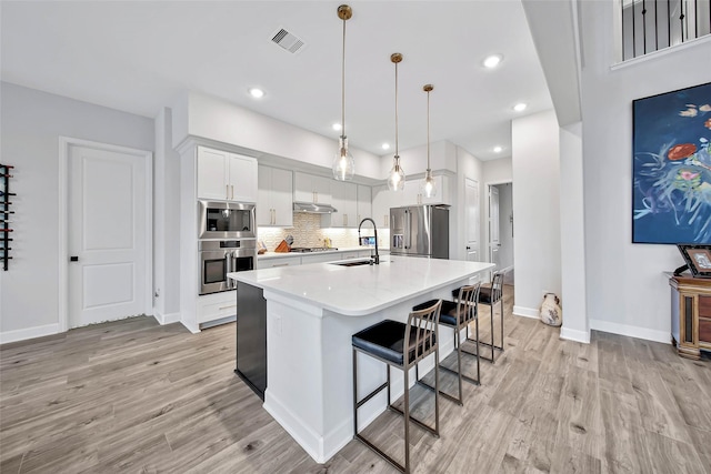 kitchen with sink, hanging light fixtures, stainless steel appliances, white cabinets, and a center island with sink