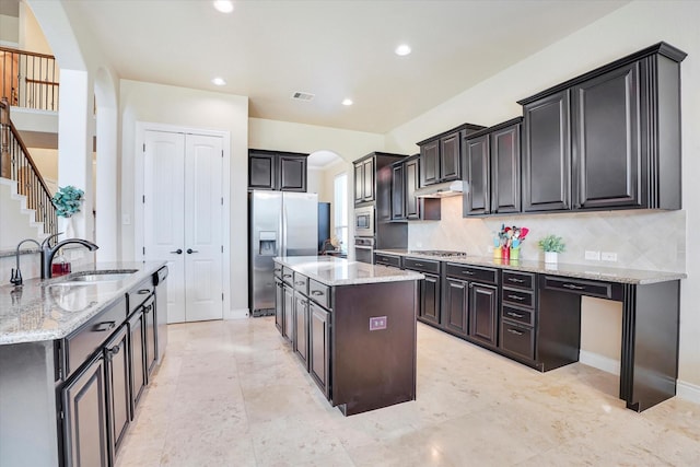 kitchen with sink, tasteful backsplash, light stone counters, appliances with stainless steel finishes, and an island with sink