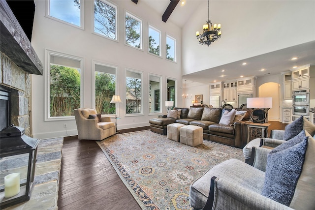 living room featuring dark wood-type flooring, a chandelier, a high ceiling, a stone fireplace, and beamed ceiling