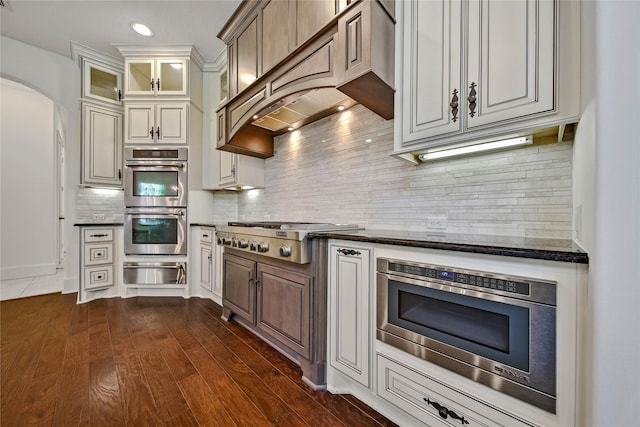 kitchen with custom exhaust hood, tasteful backsplash, dark stone countertops, appliances with stainless steel finishes, and dark hardwood / wood-style flooring