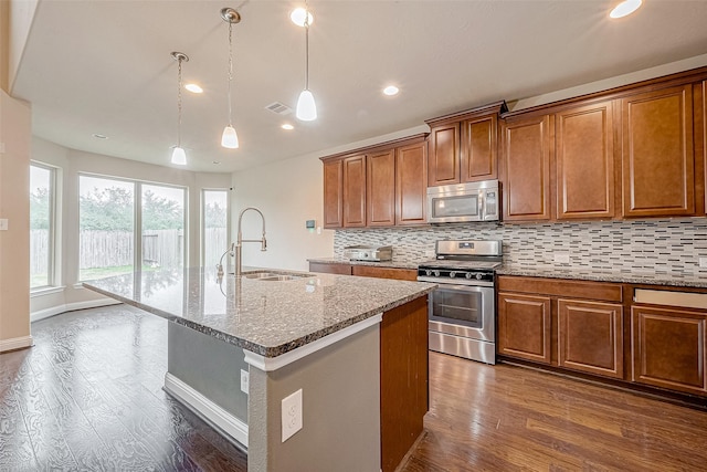 kitchen featuring dark wood-type flooring, sink, a center island with sink, appliances with stainless steel finishes, and light stone countertops