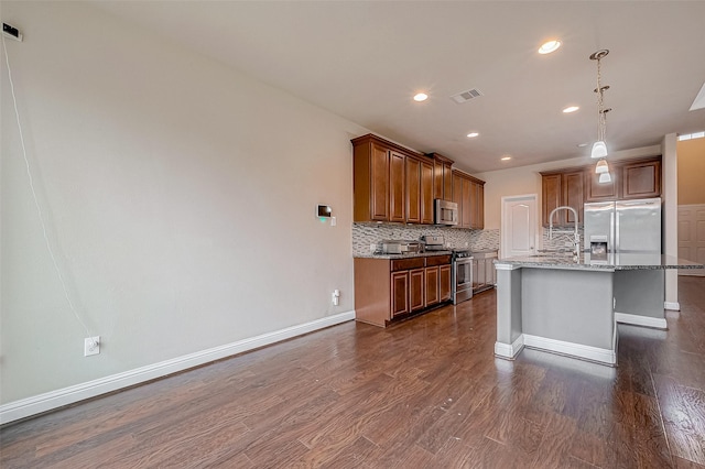 kitchen featuring a kitchen island with sink, backsplash, stainless steel appliances, light stone countertops, and dark hardwood / wood-style flooring