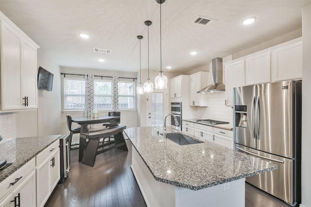 kitchen with wall chimney exhaust hood, white cabinetry, a center island with sink, appliances with stainless steel finishes, and backsplash