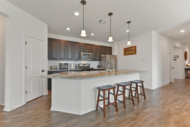 kitchen with pendant lighting, a kitchen island with sink, stainless steel appliances, dark brown cabinetry, and decorative backsplash