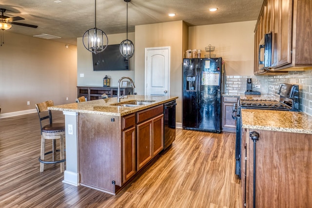 kitchen with sink, light stone counters, an island with sink, pendant lighting, and black appliances