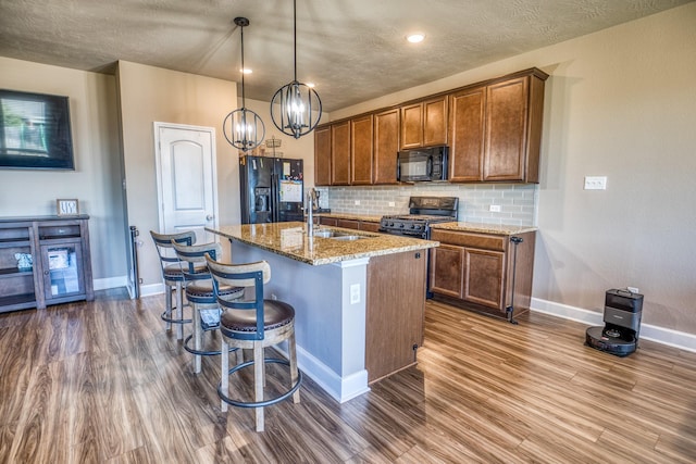 kitchen featuring sink, light stone counters, a center island with sink, pendant lighting, and black appliances