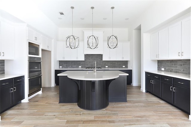 kitchen with a kitchen island with sink, double oven, decorative light fixtures, and white cabinets