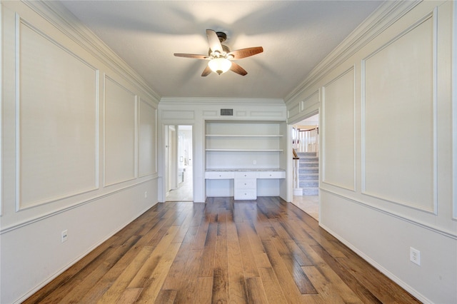 unfurnished living room with hardwood / wood-style floors, built in desk, built in features, ornamental molding, and ceiling fan