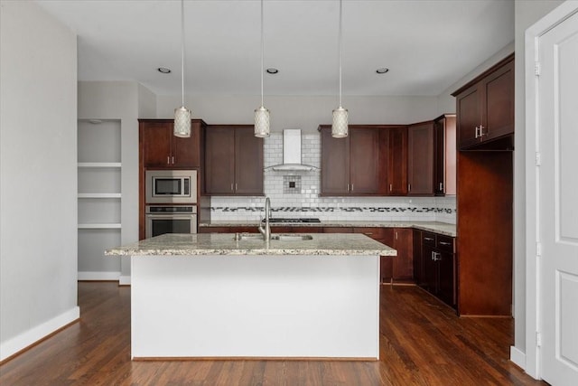 kitchen featuring wall chimney exhaust hood, light stone counters, decorative light fixtures, a center island with sink, and stainless steel appliances