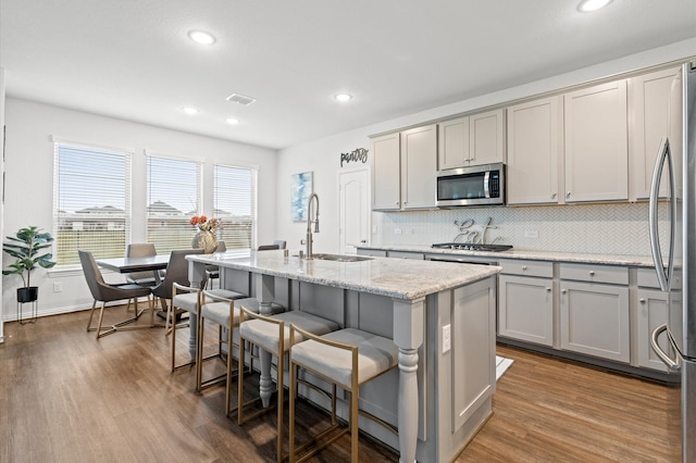 kitchen featuring appliances with stainless steel finishes, sink, gray cabinetry, light stone counters, and a center island with sink