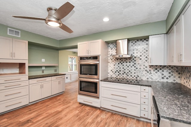 kitchen featuring wall chimney range hood, white cabinets, stainless steel double oven, and light wood-type flooring