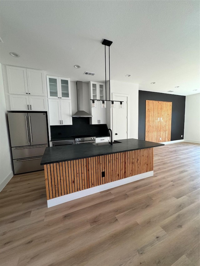 kitchen featuring wall chimney range hood, appliances with stainless steel finishes, hanging light fixtures, white cabinets, and a center island with sink