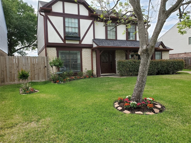 english style home featuring brick siding, a shingled roof, fence, a front yard, and stucco siding