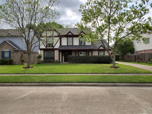 tudor house featuring a shingled roof, fence, a front lawn, and brick siding