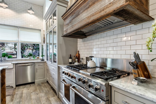 kitchen with custom exhaust hood, tasteful backsplash, light stone counters, light hardwood / wood-style flooring, and appliances with stainless steel finishes