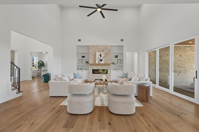 living room featuring built in shelves, a ceiling fan, stairs, light wood-type flooring, and a brick fireplace