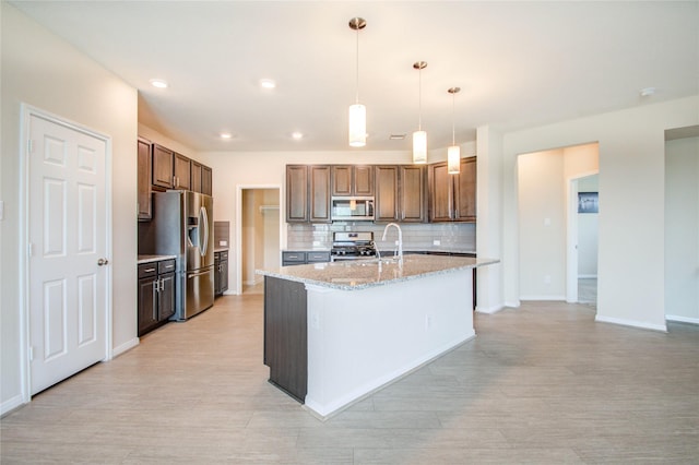 kitchen featuring hanging light fixtures, backsplash, stainless steel appliances, light stone counters, and a center island with sink