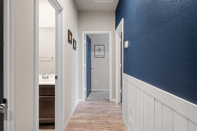 corridor with light wood finished floors, wainscoting, a sink, and visible vents