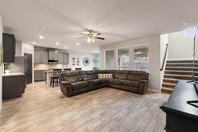 living room featuring baseboards, visible vents, light wood-style flooring, ceiling fan, and stairs