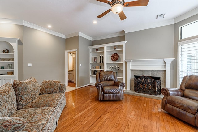 living room with built in shelves, crown molding, light hardwood / wood-style flooring, ceiling fan, and a fireplace