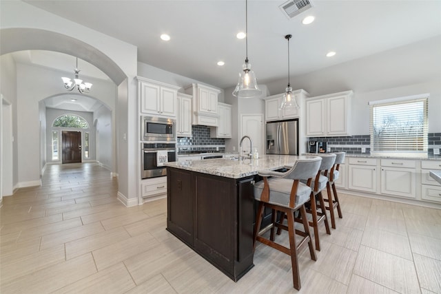 kitchen featuring white cabinetry, light stone counters, decorative light fixtures, an island with sink, and stainless steel appliances