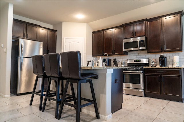 kitchen featuring a breakfast bar, backsplash, dark brown cabinets, stainless steel appliances, and light stone counters