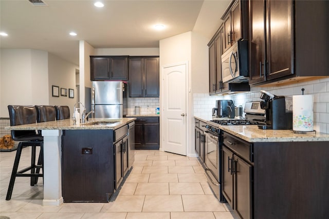 kitchen with dark brown cabinets, stainless steel appliances, a center island with sink, light stone counters, and a kitchen bar