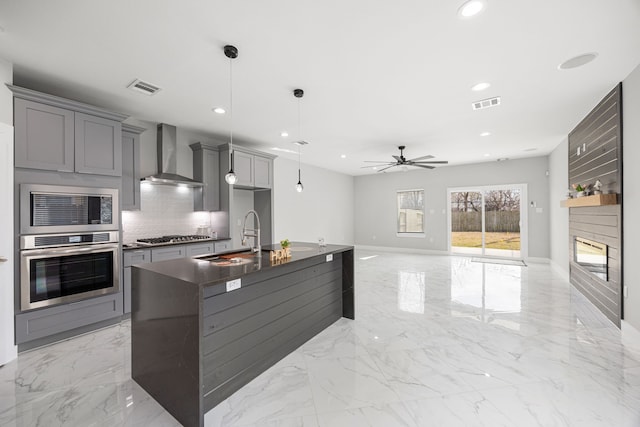 kitchen featuring wall chimney range hood, gray cabinetry, stainless steel appliances, a center island with sink, and decorative light fixtures