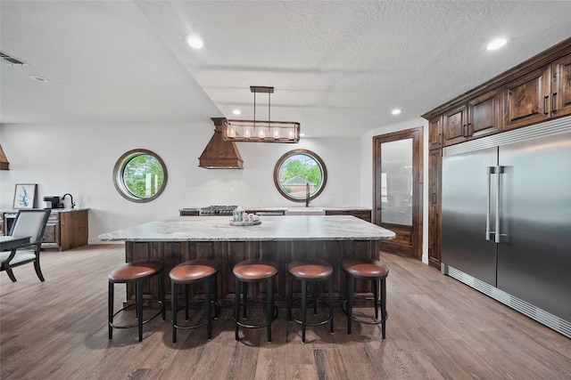 kitchen with a spacious island, a kitchen bar, decorative light fixtures, built in fridge, and light stone countertops