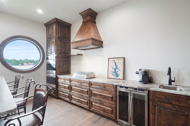 kitchen featuring dark brown cabinets, wine cooler, light stone counters, light hardwood / wood-style floors, and custom range hood