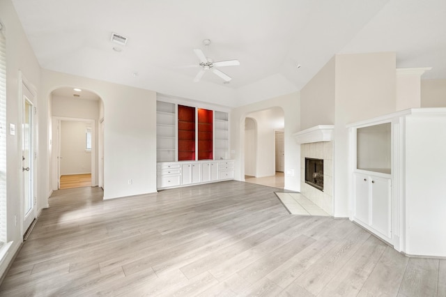 unfurnished living room with a tile fireplace, ceiling fan, light wood-type flooring, and built in shelves