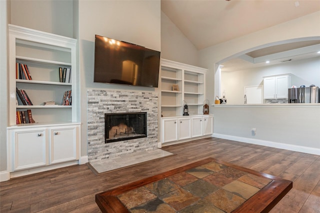 living room featuring a stone fireplace, dark wood-type flooring, built in shelves, and vaulted ceiling