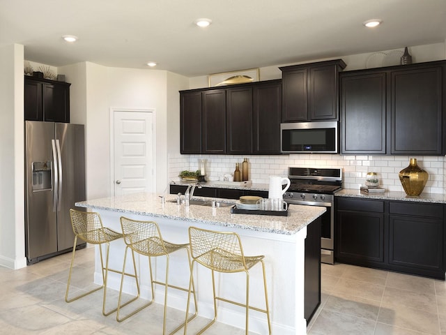 kitchen with sink, a kitchen island with sink, stainless steel appliances, light stone countertops, and decorative backsplash