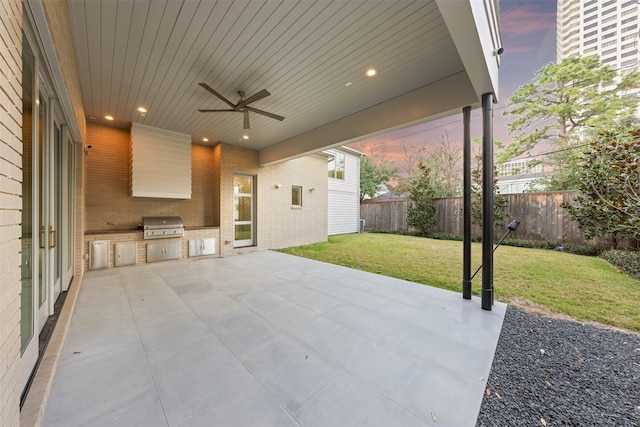 patio terrace at dusk with area for grilling, a yard, ceiling fan, and an outdoor kitchen