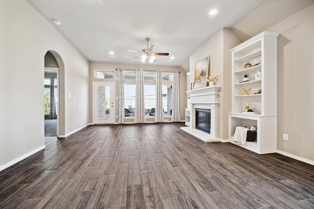 unfurnished living room with arched walkways, dark wood finished floors, a ceiling fan, a glass covered fireplace, and baseboards