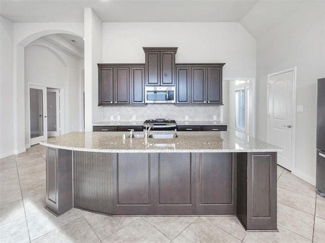 kitchen with dark brown cabinetry, stainless steel appliances, light stone countertops, and a kitchen island with sink