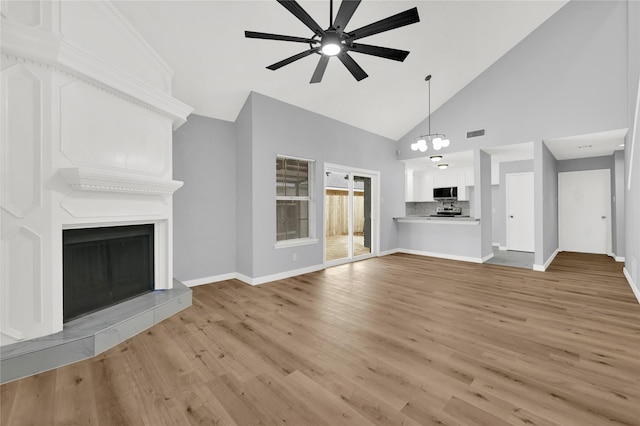 unfurnished living room featuring ceiling fan with notable chandelier, high vaulted ceiling, a fireplace, and light hardwood / wood-style floors