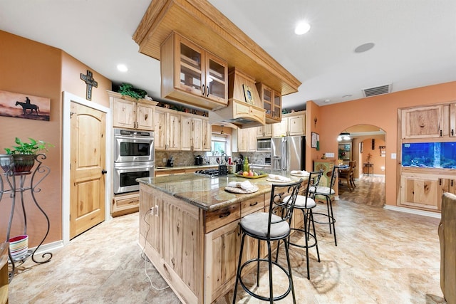 kitchen featuring light brown cabinets, appliances with stainless steel finishes, a kitchen island, light stone countertops, and decorative backsplash