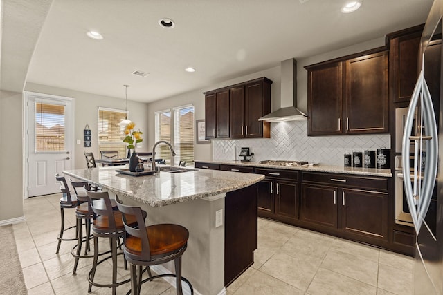 kitchen featuring appliances with stainless steel finishes, sink, hanging light fixtures, a kitchen island with sink, and wall chimney range hood