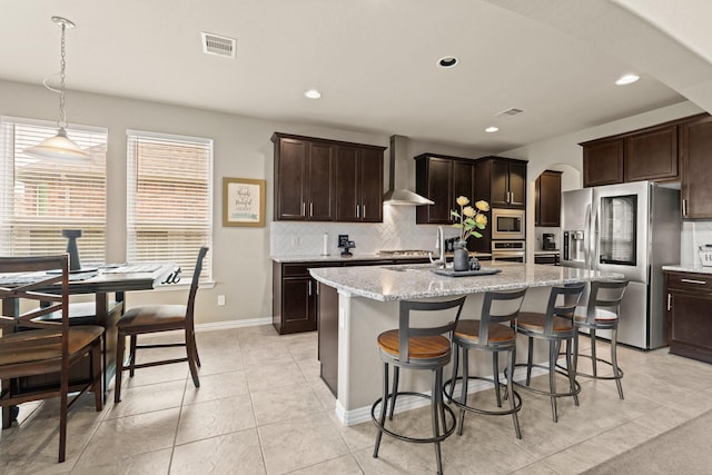 kitchen featuring a kitchen island with sink, wall chimney range hood, dark brown cabinets, and appliances with stainless steel finishes