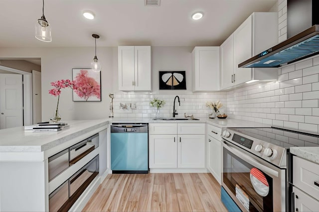 kitchen featuring a peninsula, a sink, stainless steel appliances, light wood-style floors, and wall chimney exhaust hood