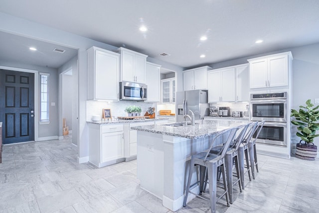 kitchen with white cabinetry, light stone counters, stainless steel appliances, and an island with sink