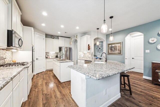 kitchen featuring appliances with stainless steel finishes, a kitchen island with sink, hanging light fixtures, white cabinetry, and light stone countertops