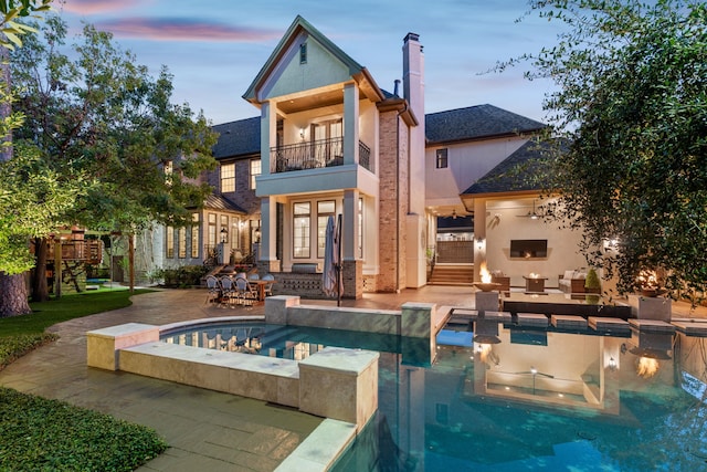 back house at dusk featuring a swimming pool with hot tub, a balcony, and a patio area