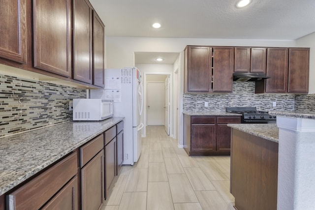 kitchen with black gas range, white microwave, light stone countertops, under cabinet range hood, and backsplash