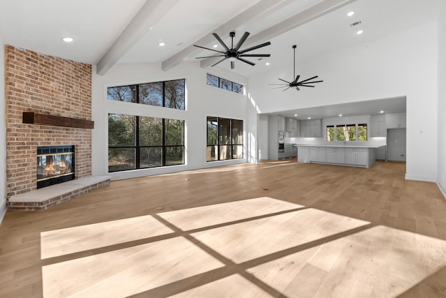 unfurnished living room featuring light wood-type flooring, beamed ceiling, ceiling fan, a fireplace, and a high ceiling