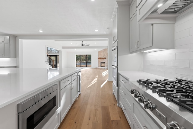 kitchen featuring appliances with stainless steel finishes, a fireplace, backsplash, custom exhaust hood, and light hardwood / wood-style flooring