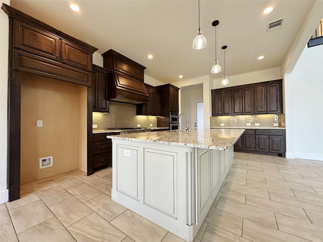 kitchen featuring pendant lighting, an island with sink, decorative backsplash, dark brown cabinetry, and light stone countertops