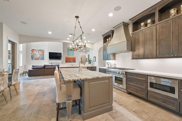 kitchen featuring appliances with stainless steel finishes, a breakfast bar, an island with sink, custom exhaust hood, and light stone countertops